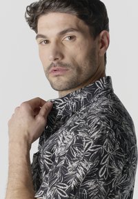 Homme aux cheveux courts et foncés ajustant le col d'une chemise noire à motif de feuilles blanches, regardant vers la caméra sur un fond uni.