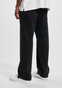 Pantalones de mezclilla negros, holgados, con un diseño de pierna recta y dos bolsillos traseros. Combinados con zapatillas blancas para crear contraste.