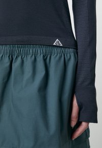 Haut noir à manches longues avec un tissu texturé, associé à une jupe verte froncée. Présente des trous pour les pouces et un logo triangulaire.