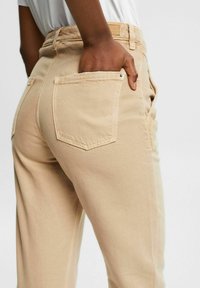Pantalons en coton beige dotés d'une taille haute, de jambes droites et de poches arrière, avec une finition texturée subtile et des détails de couture visibles.