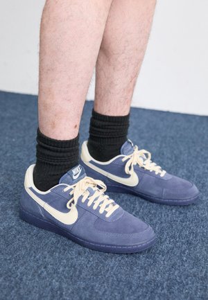 Tenisky Nike s tmavě modrým semišovým svrškem a krémovými akcenty. Obsahují ploché tkaničky, kulatou špičku a gumovou podrážku. Noseny jsou černé žebrované ponožky.