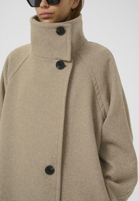 Cappotto in misto lana beige con colletto alto, caratterizzato da due bottoni neri, maniche raglan e una vestibilità rilassata. Texture liscia, design minimalista.