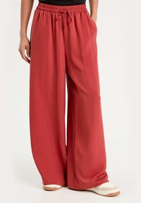 Pantalons larges dbande de couleur corail profond, avec une taille élastique et un cordon de serrage, ainsi qu'une texture côtelée. Assortis à des baskets blanches et beige.