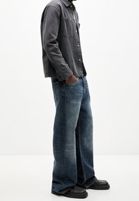 Hombre que lleva un pantalón vaquero de pierna ancha azul, zapatos negros y una camisa de botones gris oscuro, con las manos en los bolsillos, contra un fondo blanco simple.