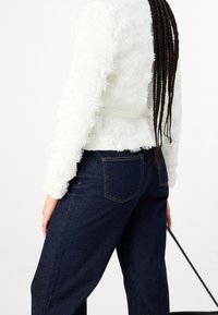 Weiße Texturjacke mit fransigen Details und einem Gürtel, kombiniert mit dunkelblauen Jeans. Das Outfit umfasst lange Zöpfe und eine schwarze Tasche.