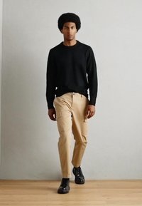 Black long-sleeve sweater, beige cargo pants with side pockets, and black shoes. Model stands on light wooden floor against a neutral wall.