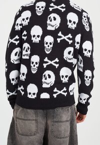 Pull noir avec motif tête de mort et os croisés, texture en tricot, ourlet et poignets côtelés, coupe décontractée, détails de couture visibles.