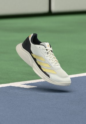 Chaussure de sport blanche et noire avec des rayures jaunes flottant au-dessus d'une surface de terrain de sport bleu et vert près d'une ligne blanche de délimitation.