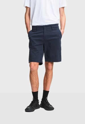 Homme portant un short bleu marine, des chaussettes noires et des baskets noires, debout les mains dans les poches devant un arrière-plan uni.
