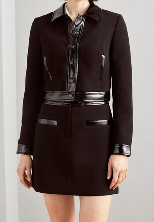 Frau trägt ein schwarzes langärmeliges Kleid mit glänzendem schwarzem Kragen, Manschetten, Knöpfen und Taschenakzenten, steht vor einer schlichten hellen Wand.