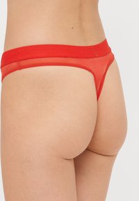 Rdeče tangice z mehkim elastičnim pasom, prosojen material, minimalna pokritost in brezšivne robove. Teksturna tkanina in živa barva.