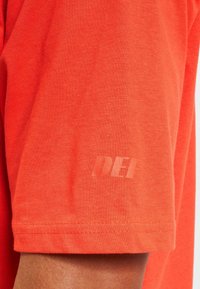 Kurzärmliges T-Shirt aus leuchtend orangefarbenem Baumwollmix, mit einem dezenten "DEF"-Druck auf dem Ärmel und einer glatten Textur.