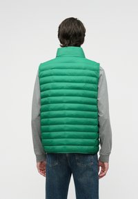 Vest verde tip puffer cu guler înalt, având un design matlasat și o textură netedă. Purtați deasupra unei cămăși gri cu mânecă lungă și blugi.