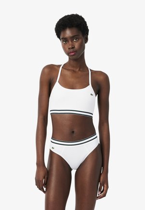 Vrouw die een witte sportbeha en slip draagt met groen-zwart gestreepte bies en klein groen logo op borst en heup, staand tegen een witte achtergrond.