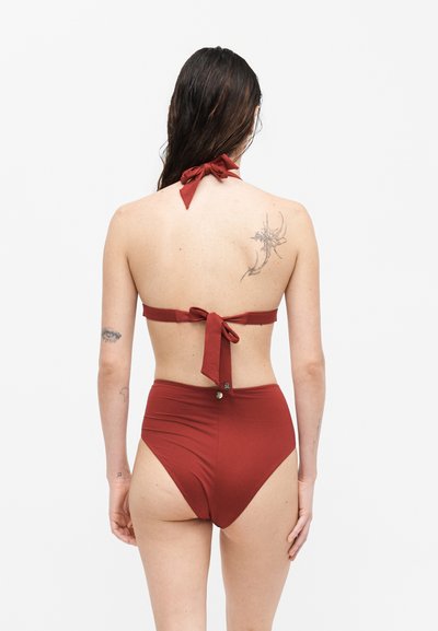 Γυναίκα με βρεγμένα μαλλιά που φορά μια κοκκινωπή διμερής μαγιό με ψηλόμεσο κάτω μέρος, εμφανίζοντας τατουάζ στην πλάτη και τα χέρια της.