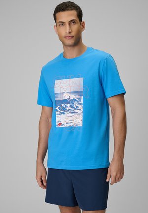 Mężczyzna ubrany w jasnoniebieski T-shirt z grafiką oceanu i napisem "SUMMER SEASON", w połączeniu z ciemnoniebieskimi szortami, stoi na jednolitym tle.