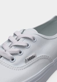 Gros plan sur une basket Vans blanche avec des lacets blancs et une étiquette en tissu affichant le logo Vans sur une tige en toile cousue.
