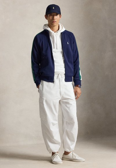 Homme portant une veste bleu marine, un sweat à capuche blanc, un pantalon cargo blanc, des baskets blanches et une casquette de baseball bleu marine, debout devant un fond uni.