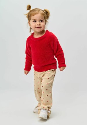 Pull rouge en peluche, pantalon en velours côtelé beige avec des motifs de cœurs rouges, et baskets blanches. Enfant avec des couettes se tient dans un espace neutre.