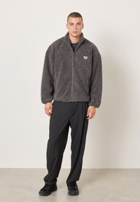 Vyras stovi dėvėdamas pilką užsegamą vilnonį švarką, juodas kelnes ir juodus sportinius batelius ant vienspalvio šviesaus fono.