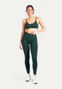 Donkergroen sportbeha en set met leggings met hoge taille, gemaakt van een gladde, rekbare stof. Model draagt witte sportschoenen en staat rechtop.