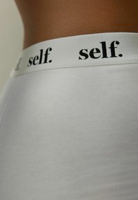 Valkoinen joustava vyötärönauha, jossa mustalla "self." -brändäys; sileä, tiukka valkoinen kangas; ribbikuviointi vaatteen pinnassa.