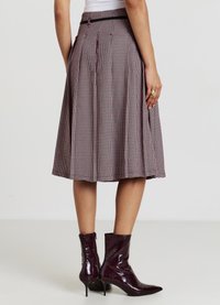 Jupe plissée taille haute à motif pied-de-poule rouge et noir, finie avec une ceinture noire, associée à des bottines bordeaux brillantes.