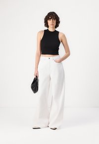 Juodas be rankovių crop top'as, baltos plačios kelnės, modelis laiko juodą rankinę. Aprangos dizainas minimalistiškas, su švariomis linijomis.