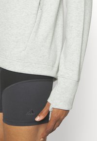 Sudadera gris claro con puños acanalados y un bolsillo con cremallera, combinado con pantalones cortos negros ajustados que cuentan con un pequeño detalle de logo. Textura suave.