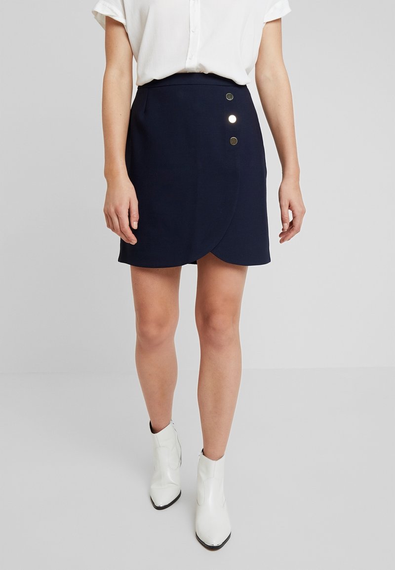 Femme portant une jupe portefeuille bleu marine à trois boutons décoratifs, une chemise blanche à manches courtes et des bottines blanches, debout sur un sol gris.
