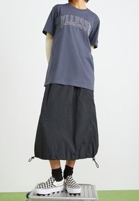Camiseta de algodón gris oscuro con el logo "ELLESSE", falda larga de nailon negra hasta el tobillo, zapatos de tela a rayas con calcetines negros en capas.