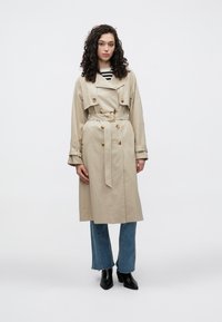 Trench-coat beige à double boutonnage, taille ceinturée et gros boutons. Porté sur un haut rayé et associé à un jean évasé bleu ainsi qu'à des bottes noires.