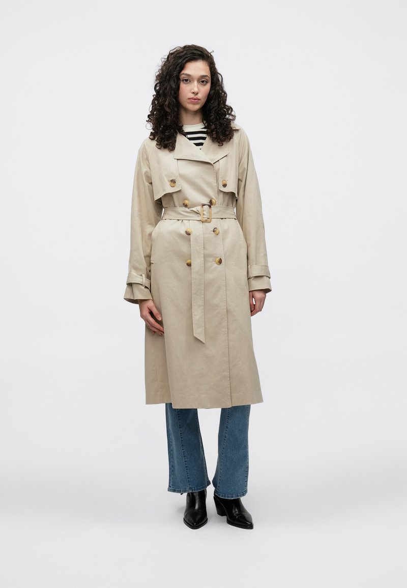 Trench-coat beige à double boutonnage, taille ceinturée et gros boutons. Porté sur un haut rayé et associé à un jean évasé bleu ainsi qu'à des bottes noires.