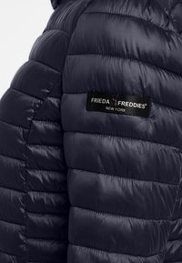 Marineblauwe pufferjack met gestikte panelen, lichtgewicht materiaal en een zwart merklabel met de tekst "FRIEDA FREDDIES NEW YORK" op de schouder.
