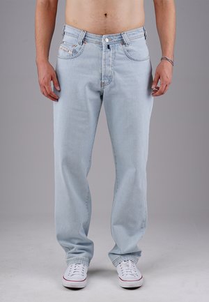 Man stands facing forward wearing light blue loose-fit jeans and white sneakers, shirtless against a neutral gray background.