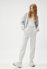 Joven con sudadera gris claro, camiseta sin mangas blanca, pantalones de chándal grises y zapatillas blancas de pie con las manos en los bolsillos frente a un fondo liso.