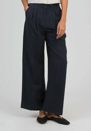 Pantalon large bleu marine à fines rayures blanches, porté avec des chaussures plates noires, mains dans les poches, sur un fond gris clair uni.