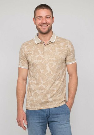 Homme souriant aux cheveux courts portant un polo beige à motifs floraux et un jean bleu, debout avec une main dans la poche devant un fond uni.