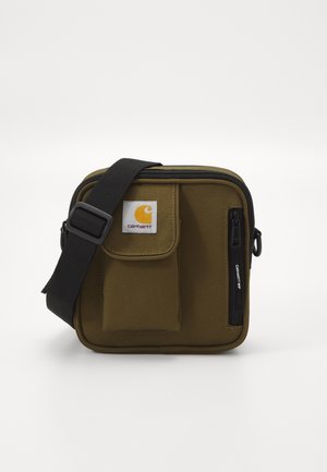 Kompaktní olivově zelená krosbody taška Carhartt z plátna s přední kapsou, černým nastavitelným popruhem a vertikální kapsou na zip na bílém pozadí.