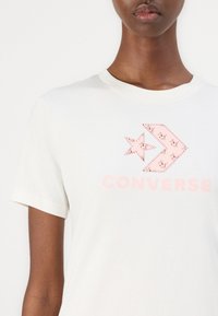 Bela bomba majica iz bombaža s roza izvezenim logotipom Converse in obliko zvezde. Kratki rokavi in krožni izrez.