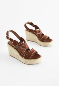 Sandali con zeppa in suede marrone, caratterizzati da cinghie incrociate con occhielli in metallo, una piattaforma in juta intrecciata naturale e una cinghia alla caviglia regolabile.