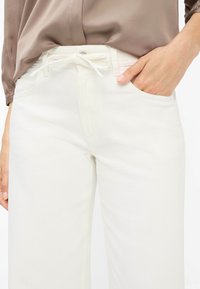 Pantalon en denim blanc avec une texture lisse, coupe droite, ceinture à cordon de serrage et poches latérales. Le matériau semble robuste tout en étant flexible.