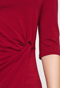 Rotes kurzärmeliges Hemd mit einem gerafften Twist-Detail an der Taille und sichtbaren Nähten am Ärmelabschluss.