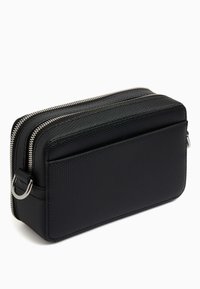 Sac rectangulaire noir en matériau texturé avec deux compartiments zippés et une boucle sur le côté pour attache. Coutures lisses et design minimaliste.
