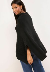 Poncho nero lavorato a maglia con spacco laterale, caratterizzato da un design a trecce testurizzate e un colletto alto. Morbido, vestibilità comoda. Indossato sopra jeans blu.