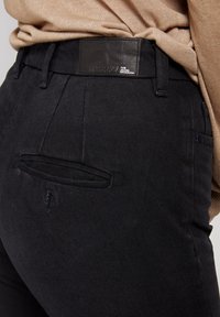 Jeans in denim nero con una texture liscia, caratterizzati da un'etichetta in pelle sulla vita e un piccolo dettaglio sulla tasca posteriore.