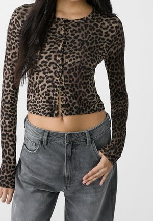Vrouw draagt een bruin-zwarte cardigan met luipaardprint en knopen en losse grijze jeans met één hand in de zak.