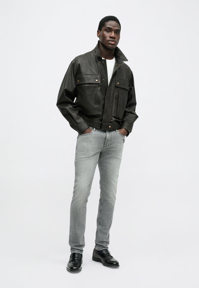 Μαύρο δερμάτινο μπουφάν με ψηλό γιακά, δύο τσέπες στο στήθος και μπροστινό φερμουάρ. Συνδυάζεται με ανοιχτό γκρι τζιν και μαύρα παπούτσια. Απλός σχεδιασμός.