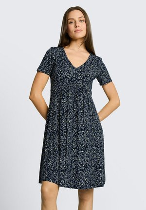 Vrouw staat met handen achter de rug, draagt een marineblauwe jurk tot op de knie met wit en geel bloemenpatroon, korte mouwen en een V-hals.