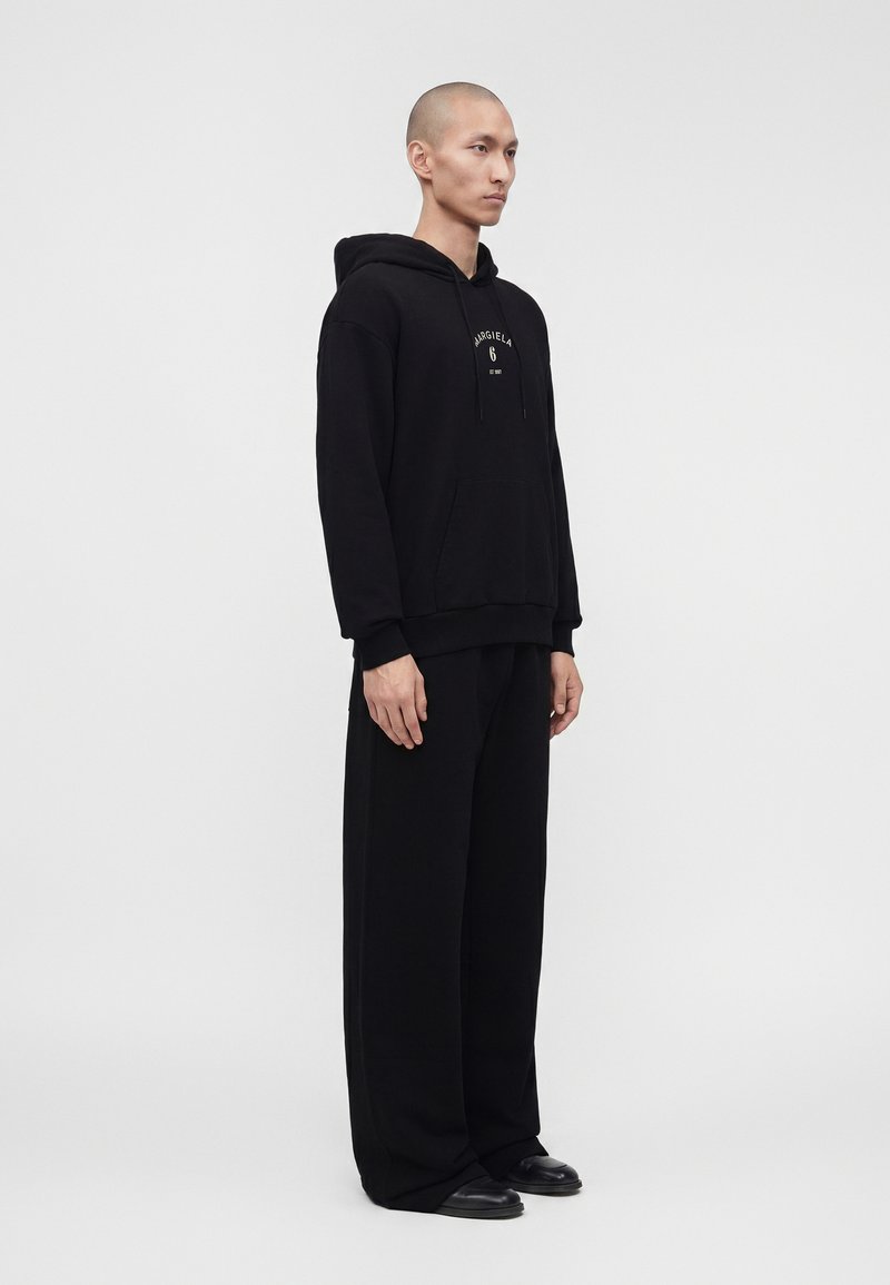 Man in een zwarte hoodie met de tekst "Margiela 6", brede zwarte broek en zwarte schoenen, zijwaarts staand tegen een effen witte achtergrond.